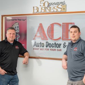 Two men dressed in a black and grey polo in front of their company sign